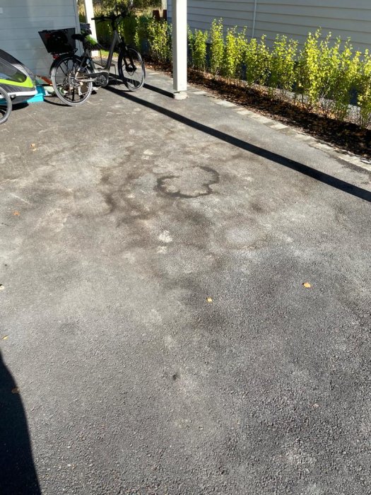 Asfalterad carport med brungråa fläckar och ringar på ytan, ett cykelparkering och häck i bakgrunden.