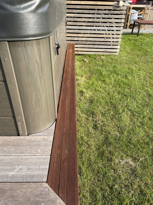 Del av en uteplats med gråa trallbrädor och en brun avslutningslist i trä vid kanten, nära en badtunna och trästaket.