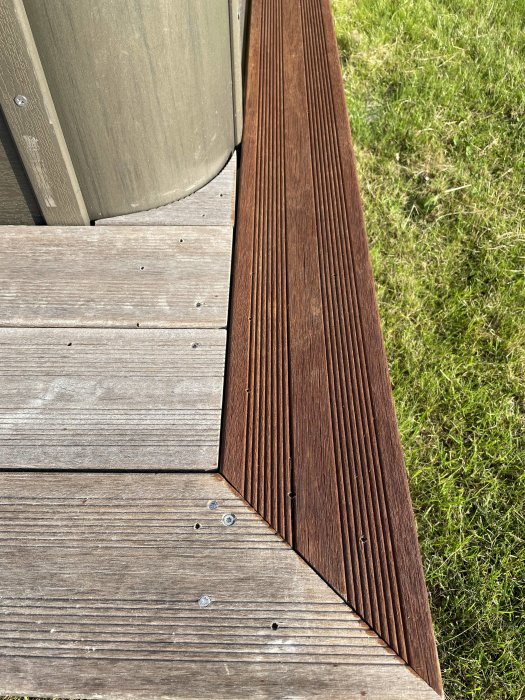 Del av en däckkonstruktion med gråa och nyanserade bruna träplankor bredvid gräsmatta i dagsljus.