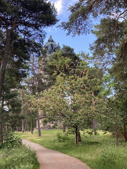 En slingrande stig genom en grönskande park med träd, leder fram mot en skulptur framför en byggnad i bakgrunden.