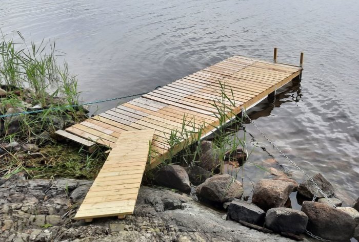 Nybyggd smal flytbrygga med landgång av trä ansluten till stenig strand och säkrad med kedja.