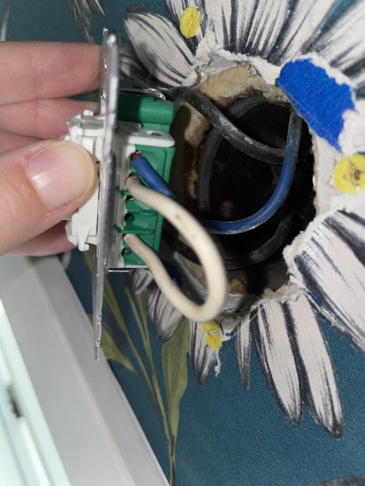 Öppen eluttag med synliga trådar, en vit kort sladd är inkopplad, mot en blommig tapetbakgrund.