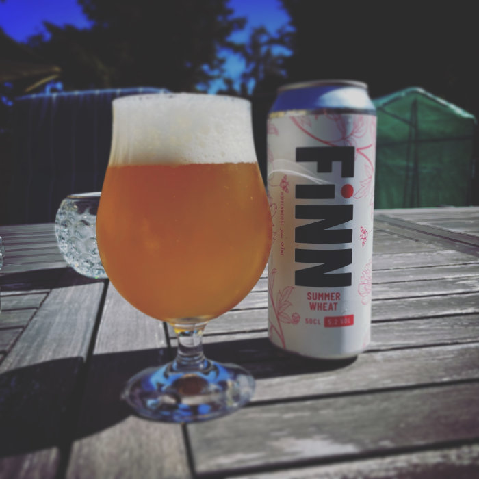 Ölglas fyllt med skummande öl bredvid en ölburk märkt FINN Summer Wheat på ett träbord utomhus.