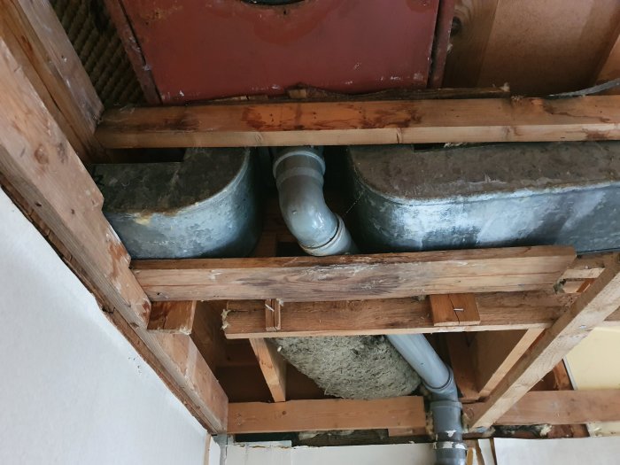 Rostig gammal ventilationstrumma som är ansluten till avloppsrör i ett orenoverat hus från 1959, synlig genom takbjälkar.