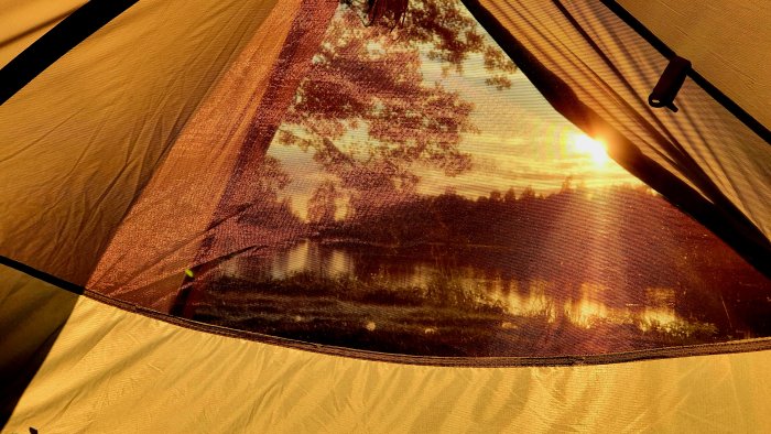 Soluppgång sedd genom en tältöppning med skog och sjö i bakgrunden.