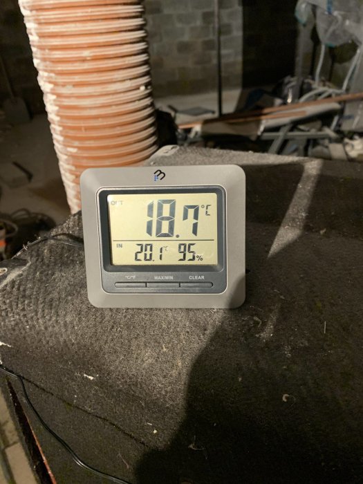 Hygrometer visar 18.7°C och 20.1% luftfuktighet i en torr källare med byggmaterial i bakgrunden.
