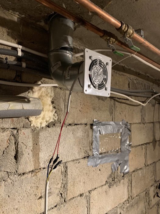 Ventilationsfläkt monterad på vägg i källare med rörledningar och isolering.