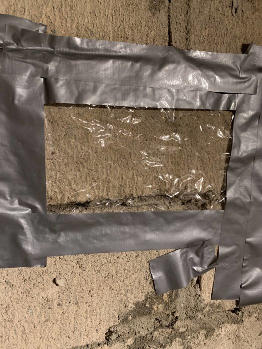 Källarvägg med silvertejp i ett rutmönster över en plastfolie som täcker ett eventuellt utrymme.