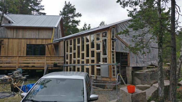 Växthus under konstruktion, ihopbyggt med hus vid en bergsklack omgivet av skog och verktyg spridda runtom.