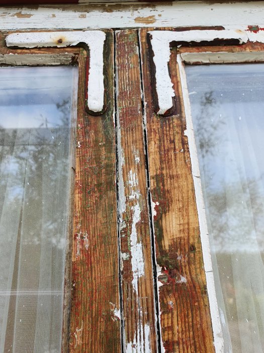 Närbild av ett gammalt fönster med genomrostade hörnjärn och sprucket kitt, samt avskalad flerfärgad målning på trä.