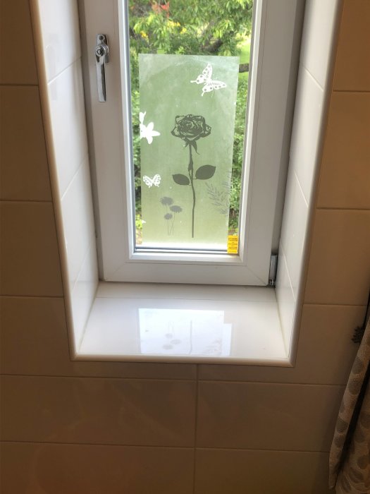 Ett badrumsfönster med en dekorativ fönsterfilm med motiv av ros och fjärilar.