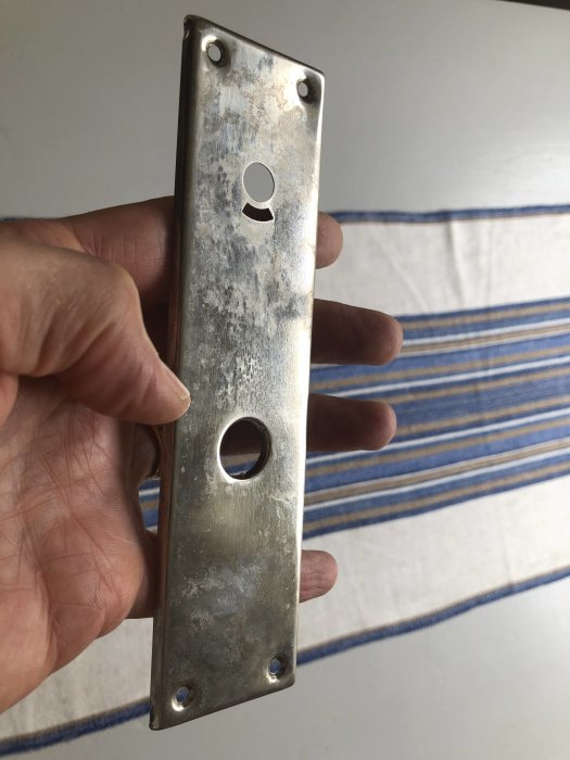 En hand håller en gammal plåtbleck dörrplatta med synlig oxidation och fläckar framför ett randigt tyg.