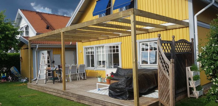 Nybyggd träpergola på däck i trädgård med husets gula fasad i bakgrunden, möbler och grönska synliga.