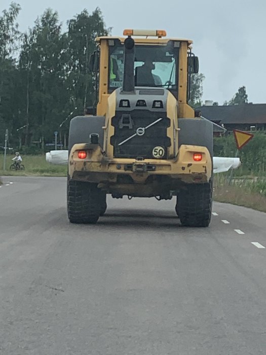 Gul hjullastare transporterar en bred markduk på en landsväg, behöver svänga ut vid möten.