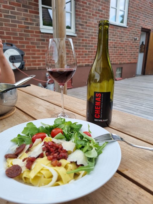 Ett glas rödvin bredvid en öppnad Beaujolais-flaska och en tallrik med pasta, sallad och tomat på ett träbord.