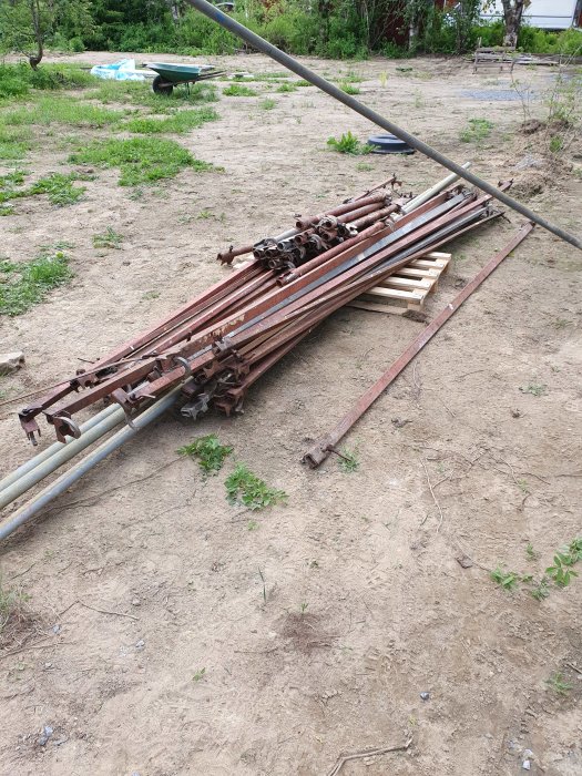 Stapel med rostiga stålrör och ställningskomponenter på en grusig mark.
