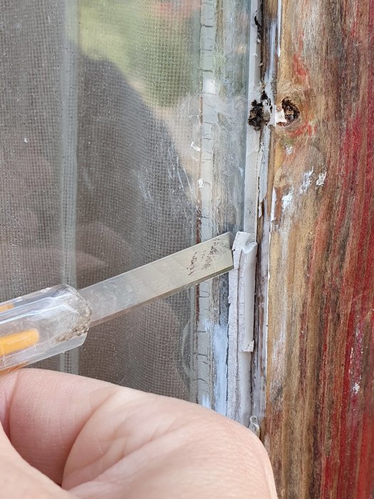 Hand som skrapar bort gammalt silikonkitt från ett fönsterhörn med en skrapa, exponerar skadade fästpunkter för hörnjärn.