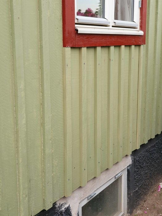 Ny grön panel under fönster med rött foder på husvägg, ny papp synlig, kalkbruk vid grund.