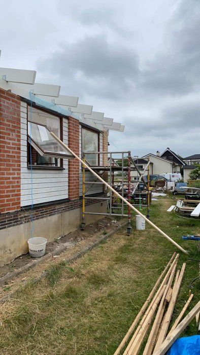 Renoveringsarbete på husfasad med ställningar, öppet fönster och byggmaterial på marken.