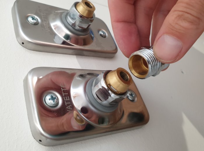 Hand som håller en rak skruvdel nära väggbrickor för VVS-installation med synliga anslutningar.