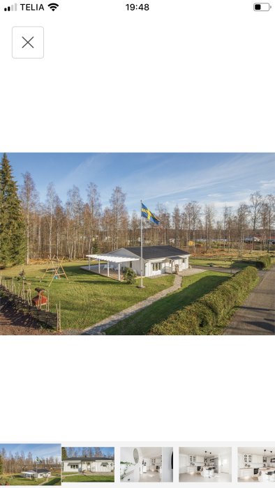 Ett enplanshus med vit fasad och svart tak med en svensk flagga, omgivet av grönt gräs och träd.