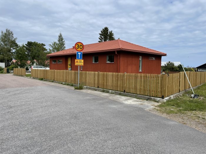 Halvfärdigt trästaket med toppade stolpar och lock, vid ett rött hus samt en gatuvy med hastighetsskylt.