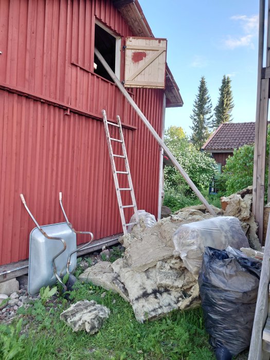 Röd stuga med öppet fönster och stege, isoleringsmaterial och trädgårdskärra på marken.