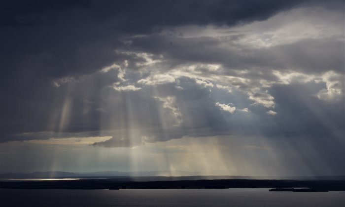 Mörka moln med solstrålar som skiner ner genom molnen över en sjö.