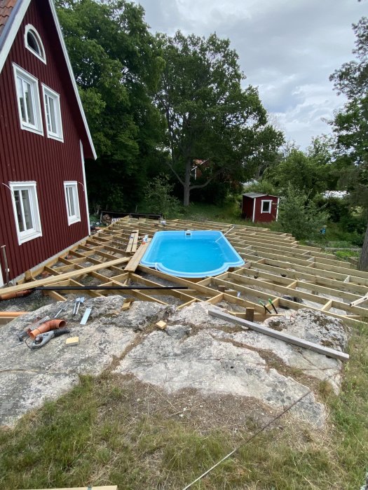 Byggstomme av trä runt en blå pool med röd stuga och verktyg i förgrunden.