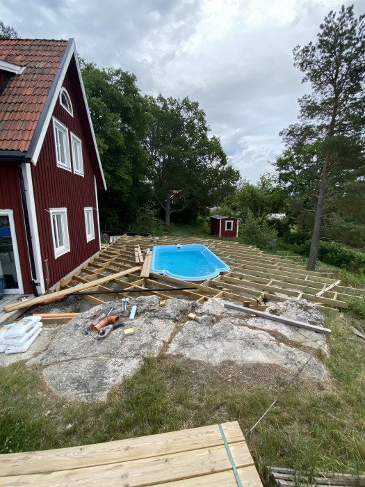 Halvfärdig trästomme för däck runt en blå pool bredvid ett rött hus, med verktyg och trämaterial.