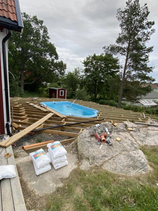 Byggstomme för däck runt ovanjordspool med byggmaterial och verktyg på gräsmatta.