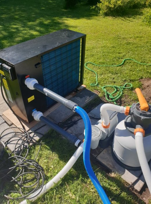 Poolvärmepump och sandfilterpump installerade på gräsmatta med anslutna slangar.