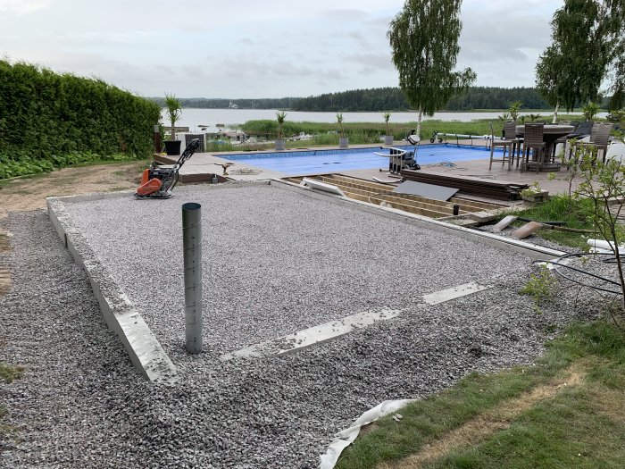 Grund till byggprojekt med mur fylld med grus och bottenprofiler framför en sjö och altan.