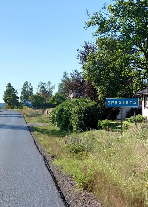 Lantväg med vägskylt som visar namnet "SPRAXKYA", grönska och del av hustak i bakgrunden.