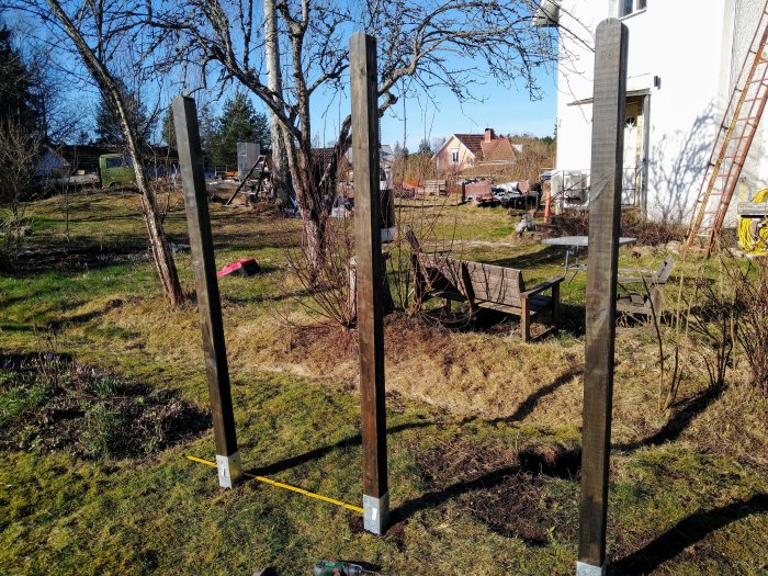 Nybyggd spaljé av trästolpar och armeringsnät i en trädgård vid en husvägg, utan klätterväxter.