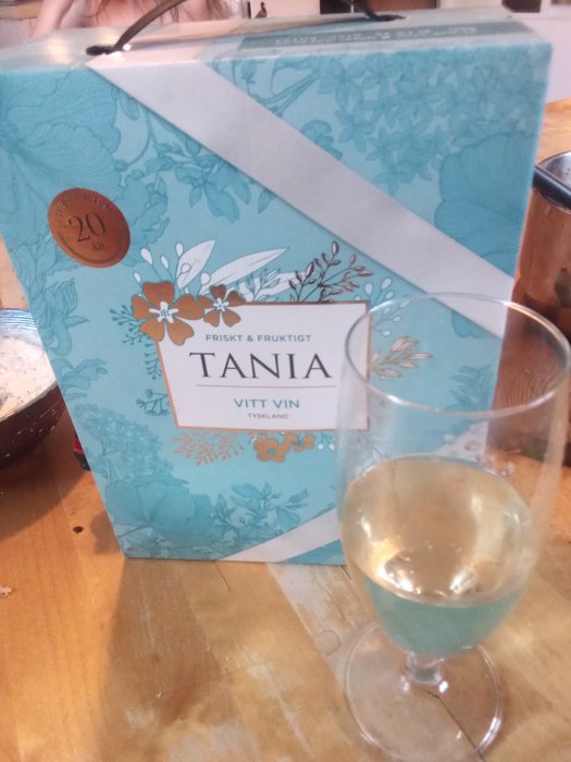 Ett ölglas på ett träbord med vitt vin bredvid en blå vinbox märkt "Tania Vitt Vin".