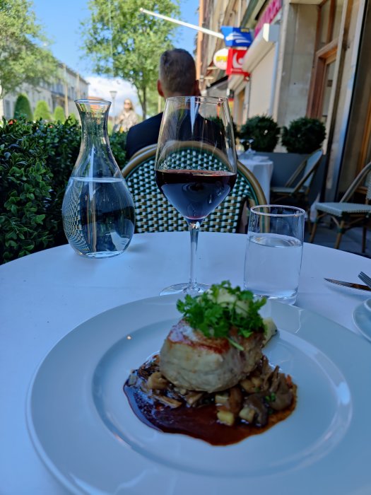 Kalvfilé med tryffelpotatis på vit tallrik, rödvinsglas och vattenkanna, utomhusrestaurang i bakgrunden.