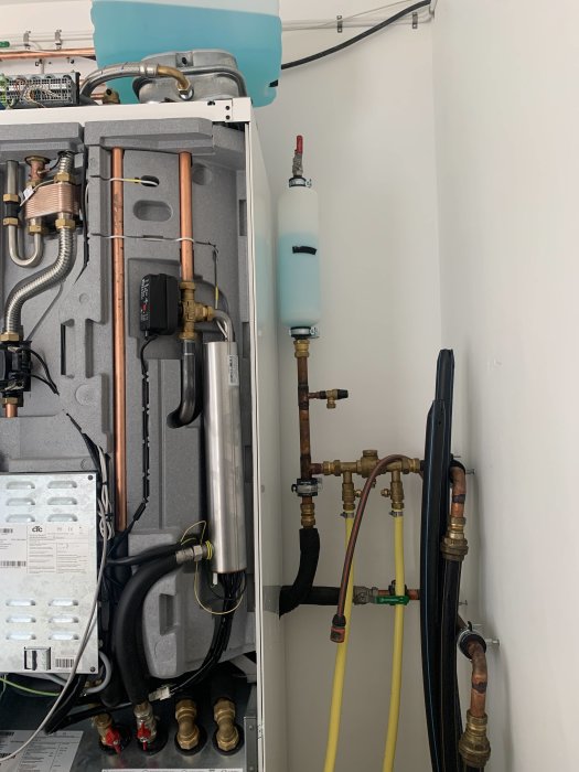 Värmepump och filtreringssystem med kopplingar, rör och IBC-tank på en vägg i ett teknikutrymme.