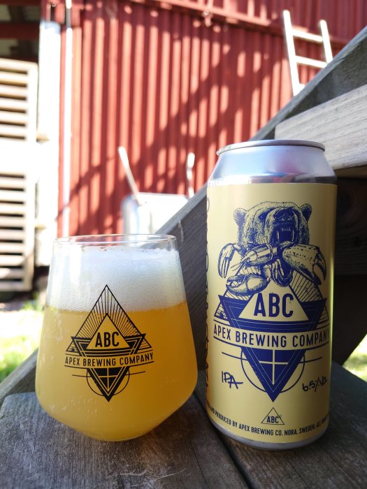 Ölglas och burk från Apex Brewing Company på en träyta med röd ladugård i bakgrunden.