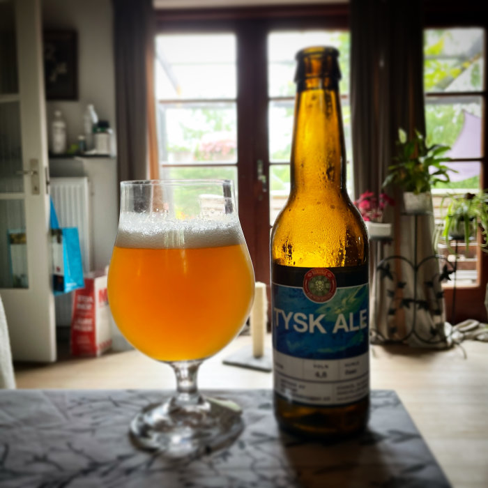 En ölglas fylld med öl intill en ölflaska med etiketten "TYSK ALE" på ett bord framför ett fönster.