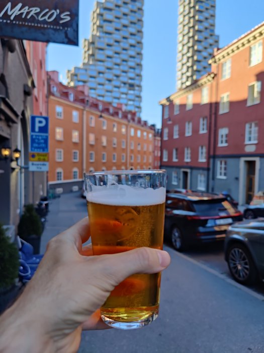 Hand håller ölglas på stadsgata med byggnader och en hög skyskrapa i bakgrunden.