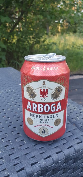 En burk Arboga Mörk Lager öl står på ett svart flätat bord med natur i bakgrunden.