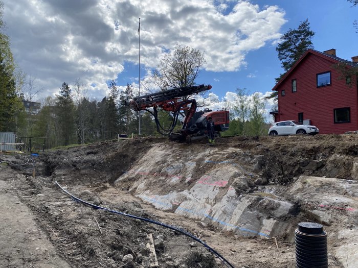 Arbetsplats med borrutrustning på sprängd tomt nära röd husbyggnad, stenar och lerig byggväg syns.