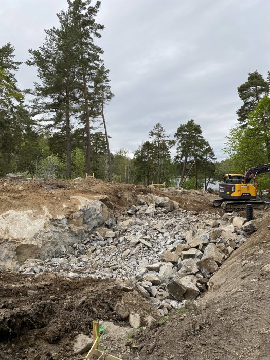 Byggarbetsplats med sprängda stenar och en grävmaskin på en tomt omgiven av träd.