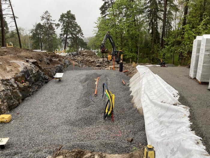 Byggplats med grusväg och dränering under arbete, grävmaskin och byggmaterial syns i bakgrunden.