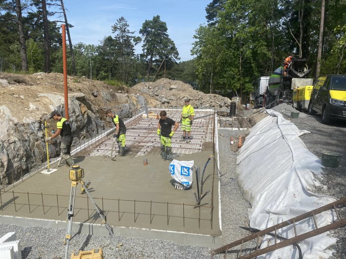 Arbetare gjuter en betongplatta med armeringsjärn på en byggarbetsplats med betongbil i bakgrunden.