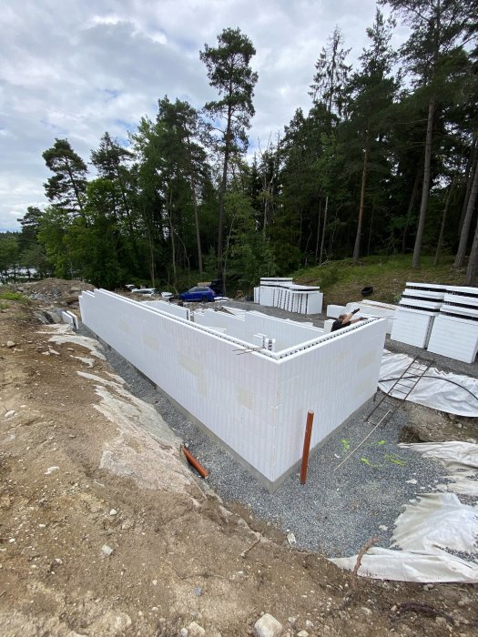 Byggarbetsplats med uppsatta vita isolerade väggelement på en betonggrund omgivet av skog.