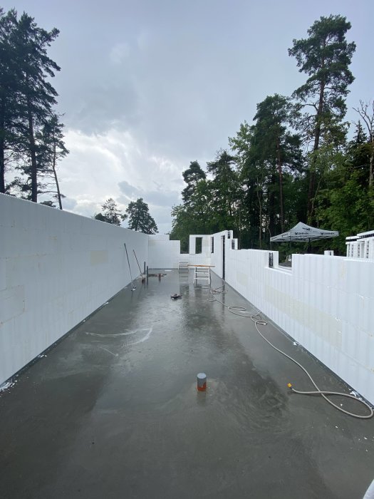 Byggplats med vita väggelement som står uppställda på en grå betongplatta, träd i bakgrunden.