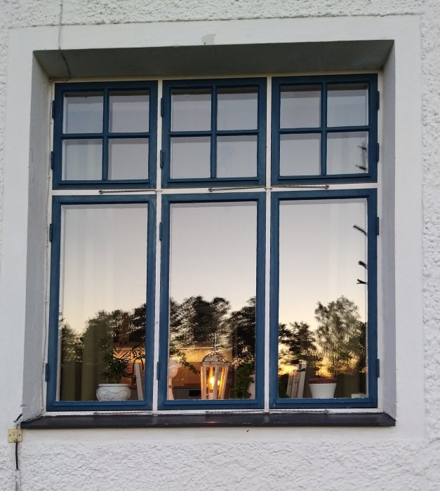 Blåmålade fönster med flaggspröjs i en vit vägg vid skymning, reflekterar interiör och solnedgång.