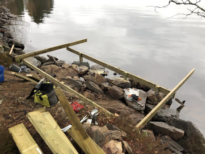 Pågående bygge av en brygga med träreglar monterade på bergig grund vid en sjö, verktyg och byggmaterial synliga.